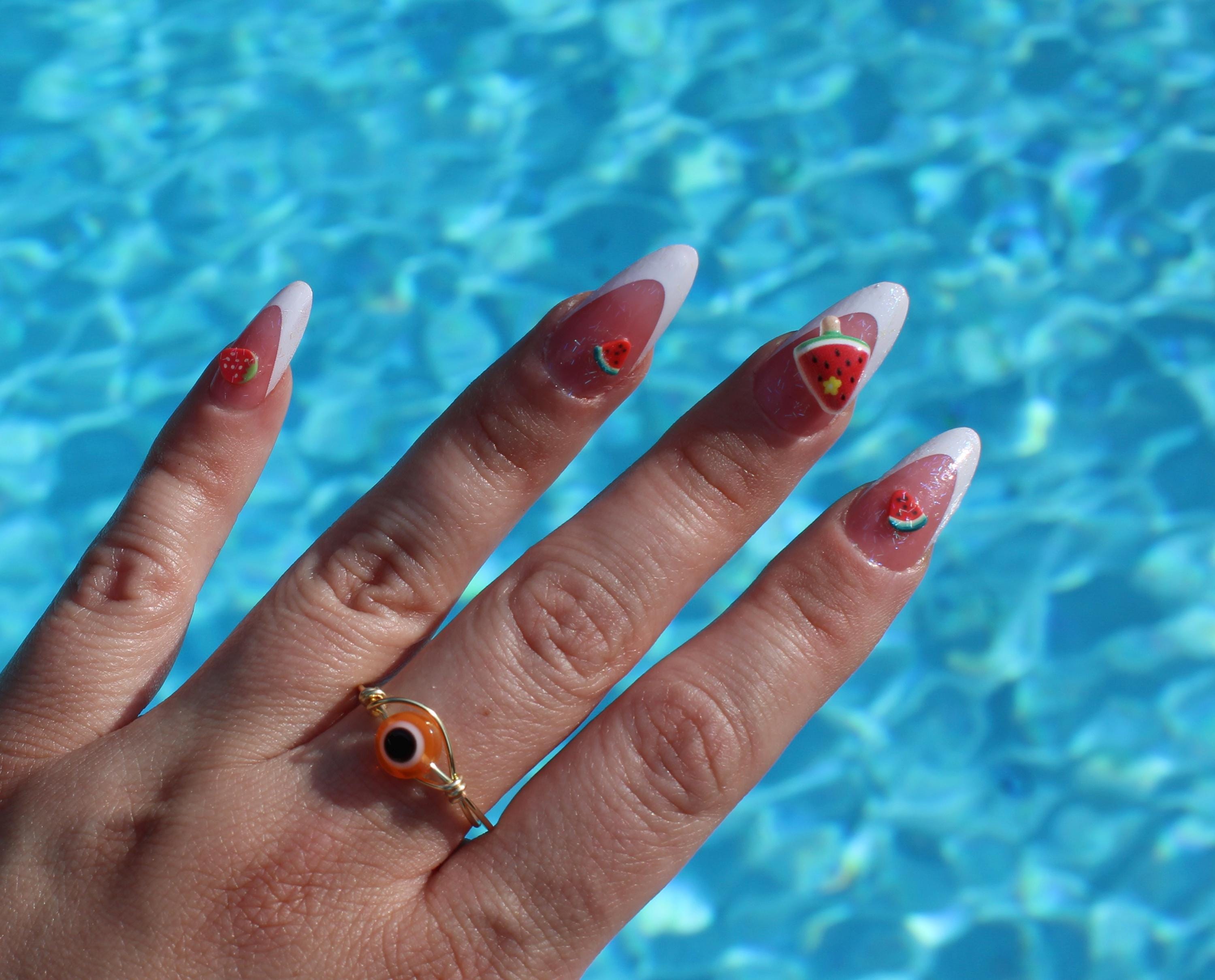 Woman's hand, featuring watermelon nail art and an orange evil eye ring, rests against a shimmering blue pool.