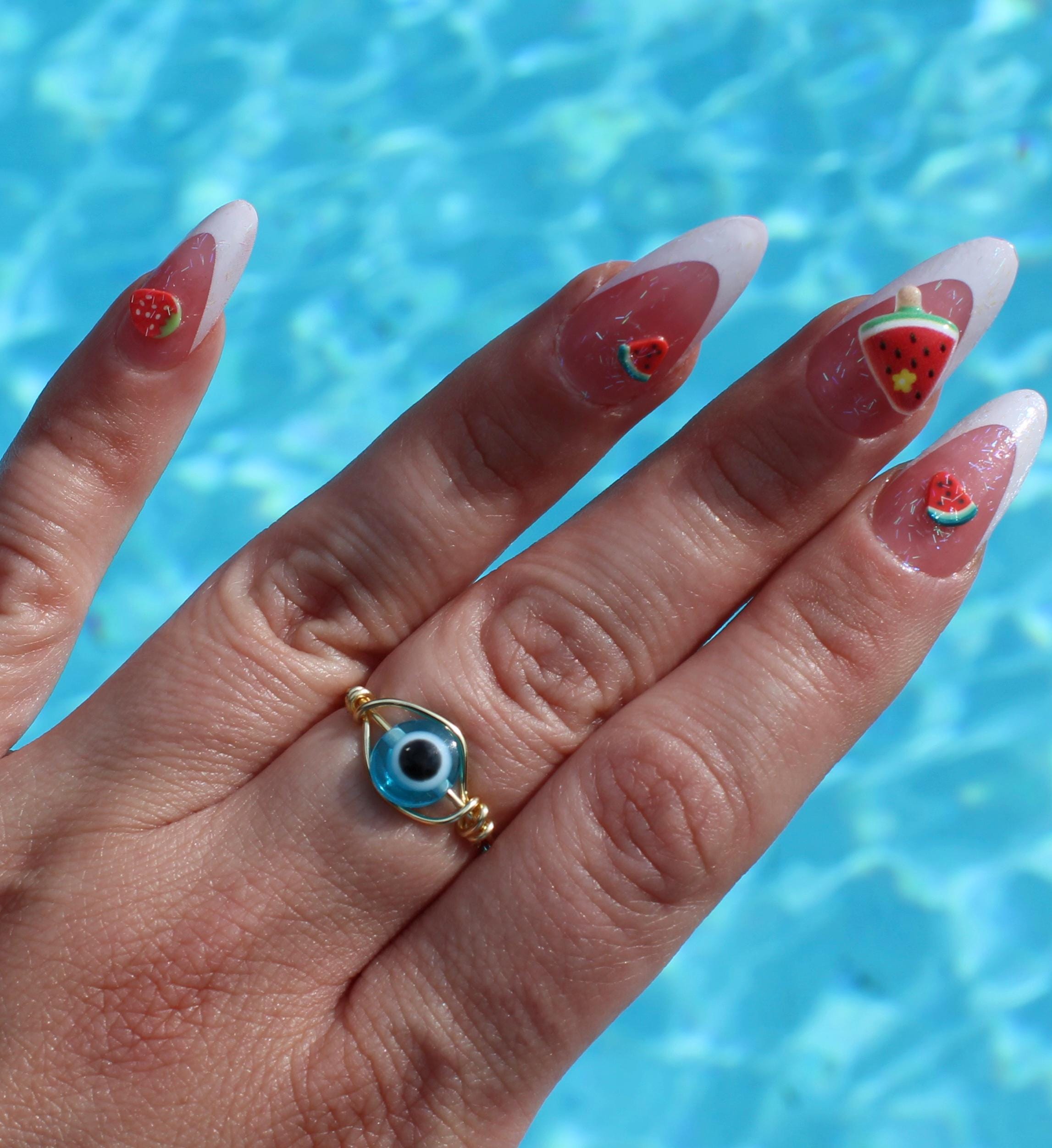 A hand models summer-themed watermelon and strawberry nail art alongside a blue evil eye ring against a pool background.