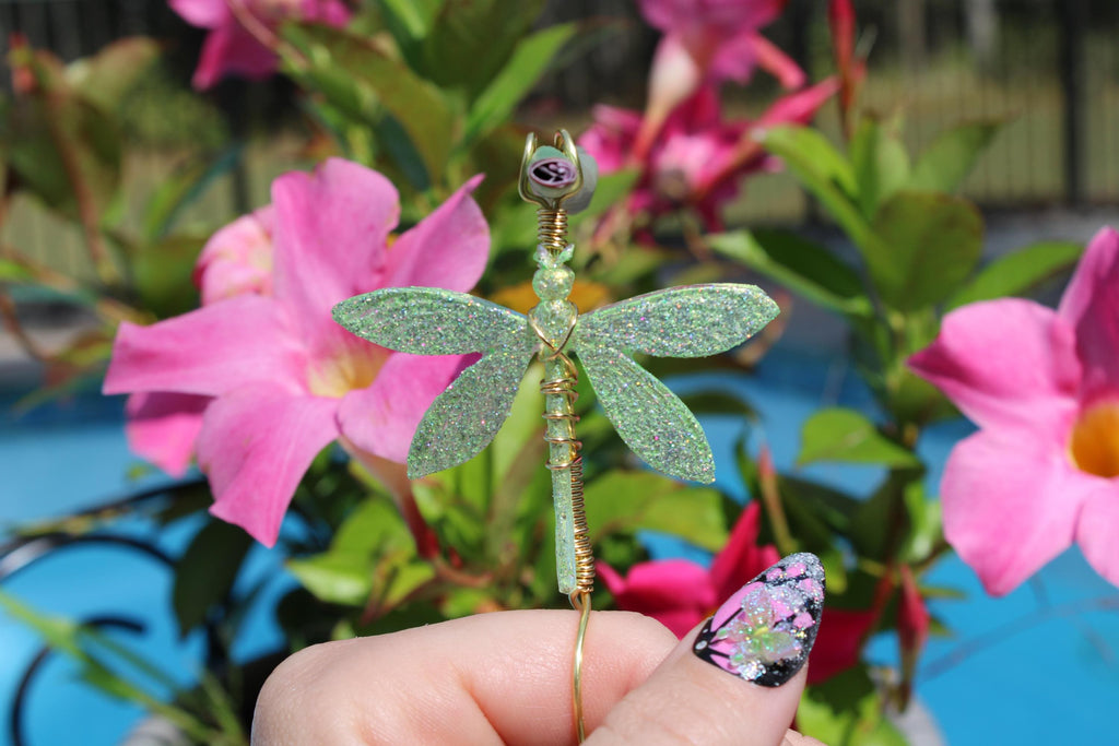 A hand holds a glittery green dragonfly smoke accessory against a backdrop of bright pink flowers.