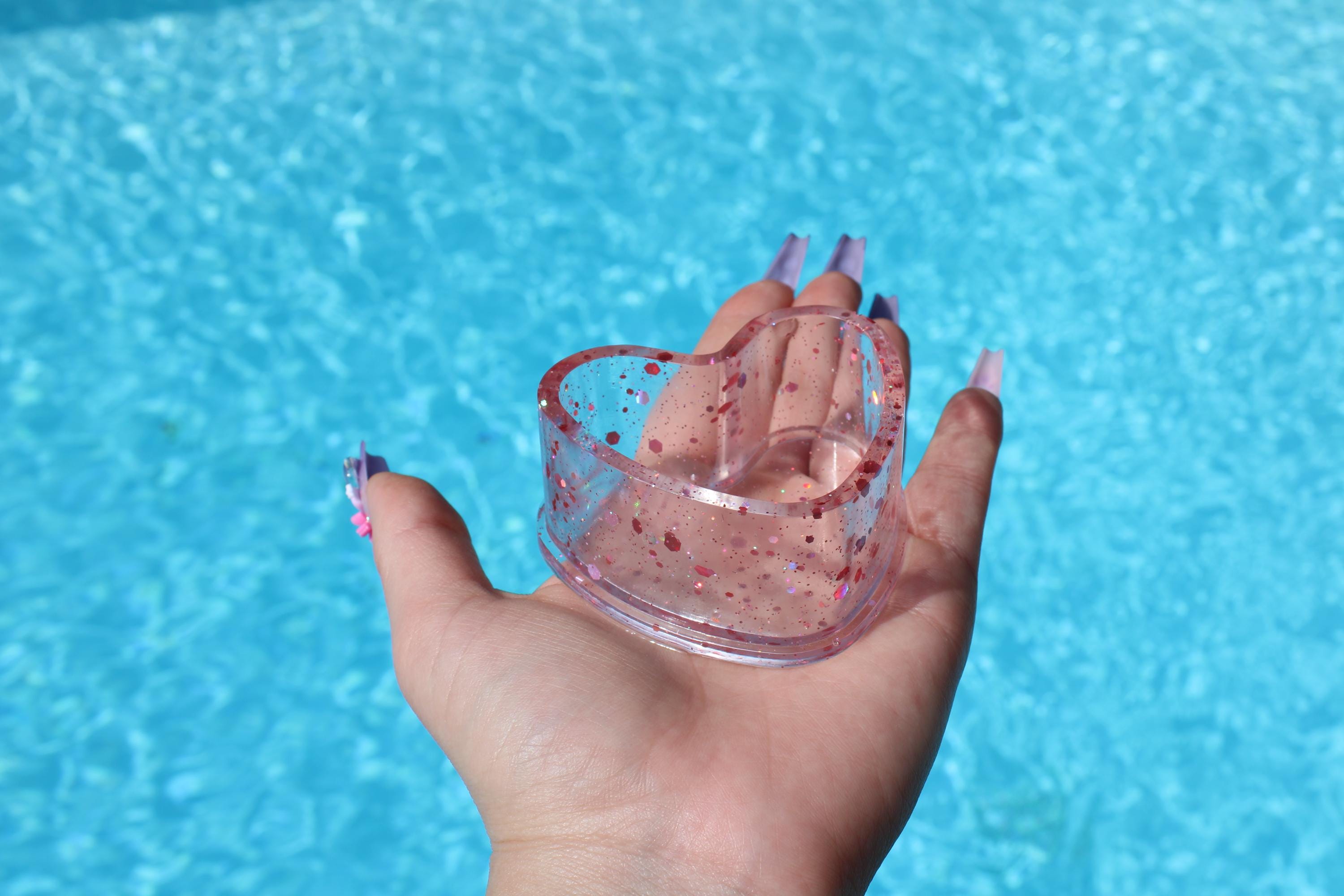 Hand holding a clear ring dish with pink specks against a blue water background