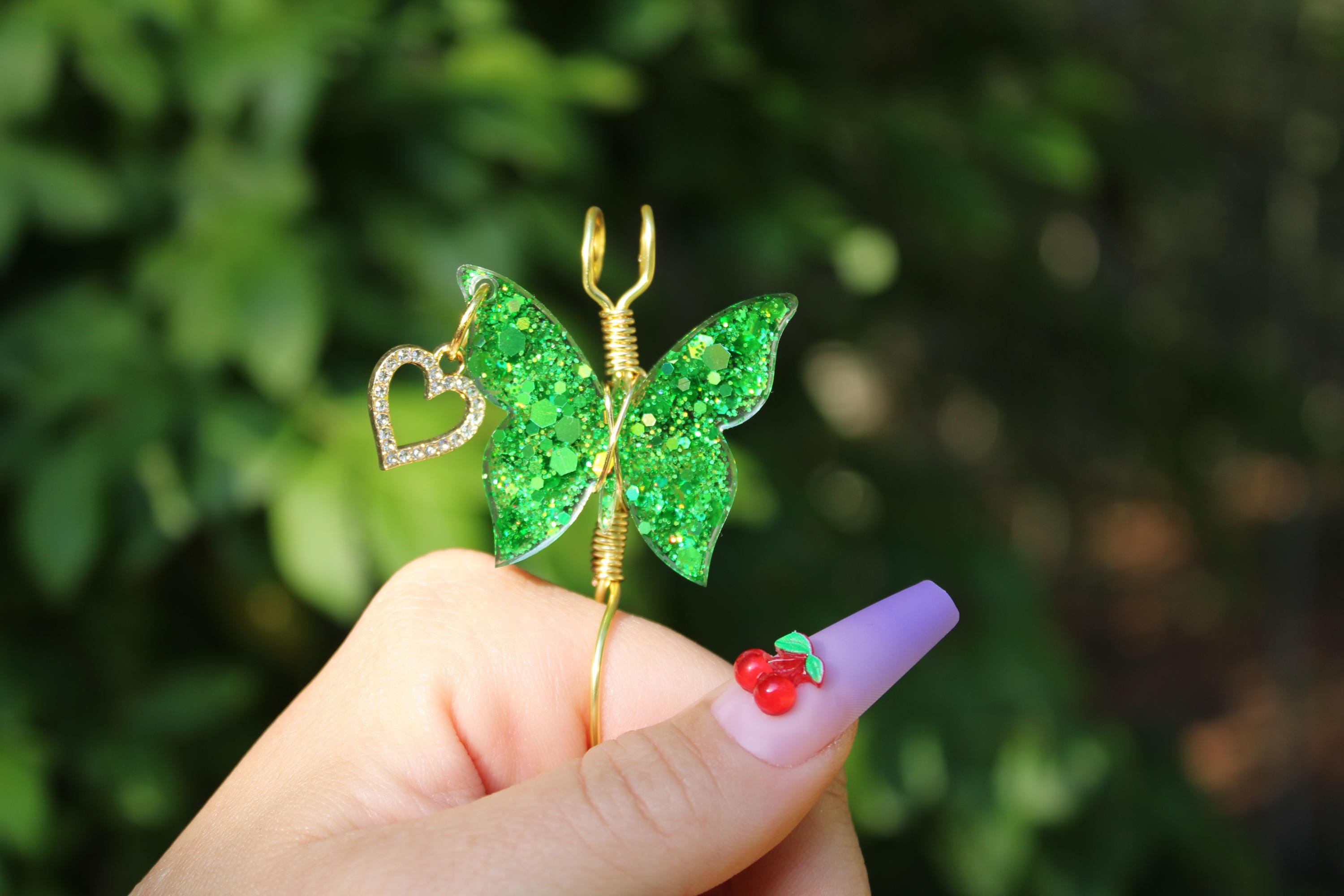 A hand displays a gold colored wire butterfly smoke ring featuring green glitter wings and a sparkling heart charm, complemented by a purple nail adorned with cherry nail art.