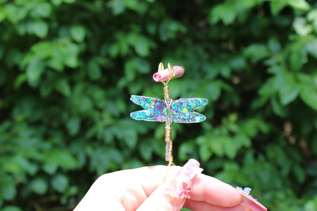 A hand with butterfly-themed nails holds a whimsical handmade dragonfly smoke ring featuring glittery turquoise wings and a pink head.