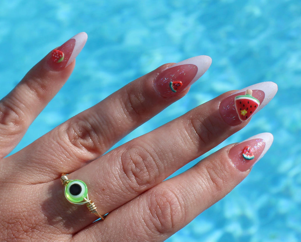 Hand with watermelon-themed nail art against a blue water background wearing a lime green evil eye ring