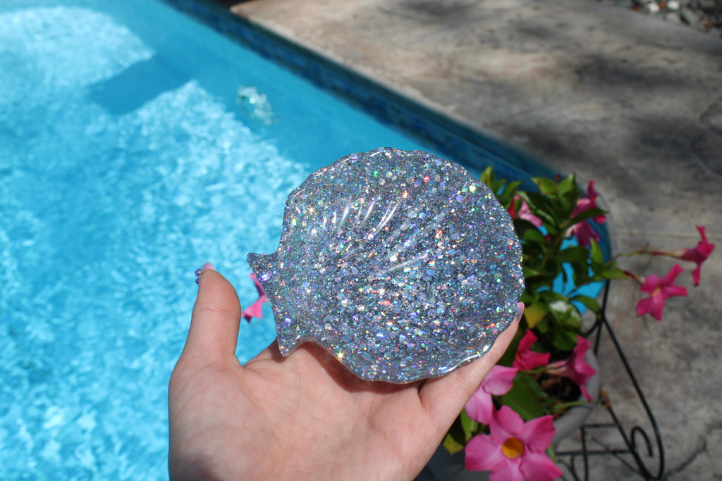Hand holding a glittery seashell soap dish near a pool with flowers in the background