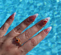 A hand showcases summer-themed nail art, featuring strawberries and watermelon, alongside an orange evil eye ring, with shimmering blue pool water as the backdrop.