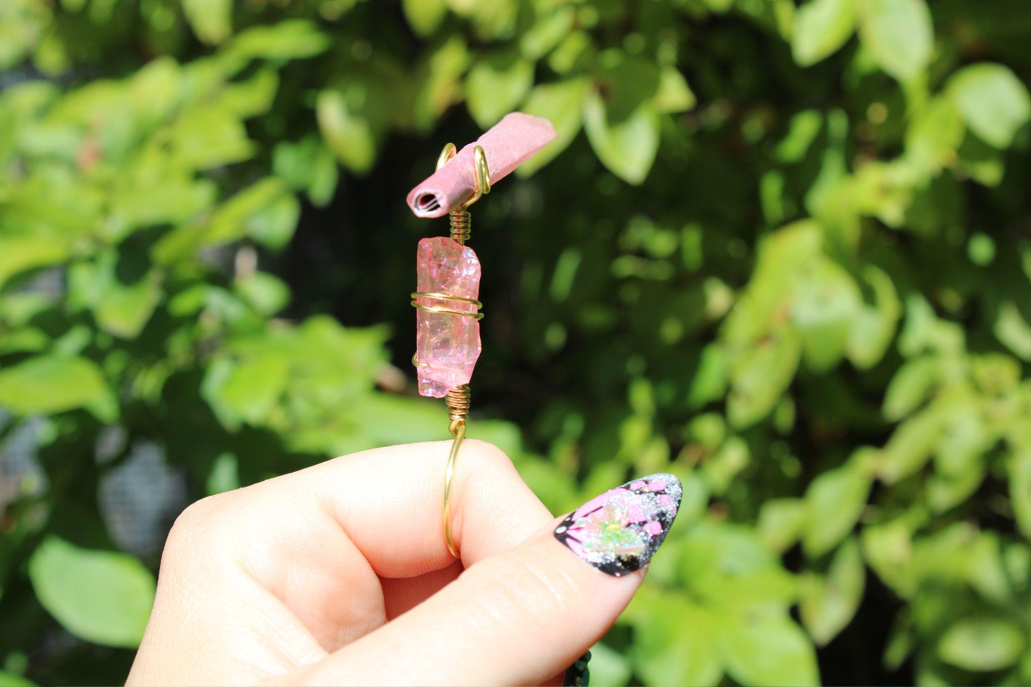 A hand with glittery nail art holds a wire-wrapped pink crystal smoke ring against a leafy green background.