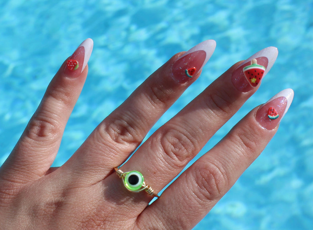Hand with watermelon-themed nail art against a pool background wearing a lime green evil eye ring