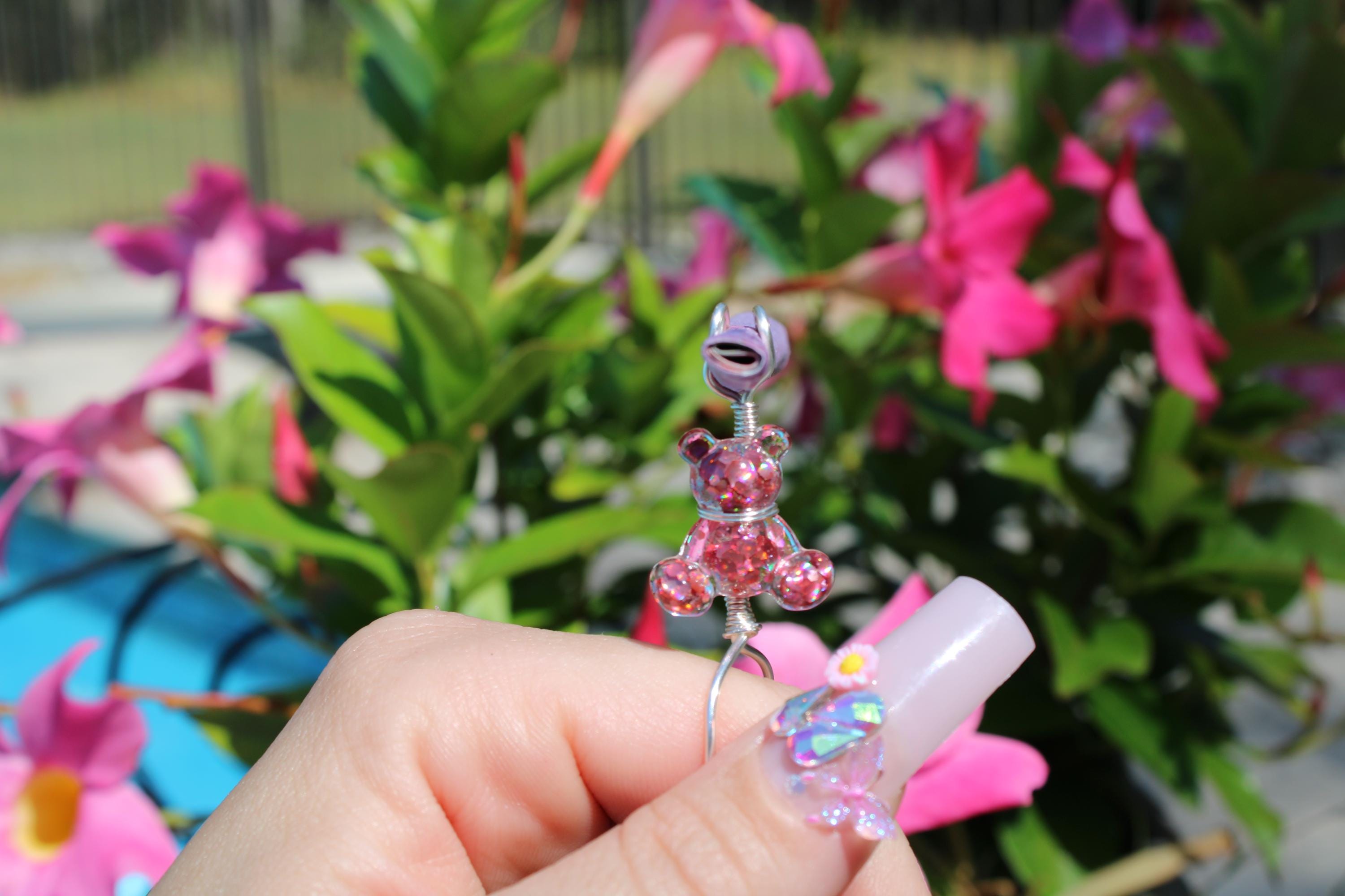 A hand with decorated nails holds a whimsical handmade wire smoke ring featuring a glittery pink teddy bear, against a backdrop of vibrant pink flowers.