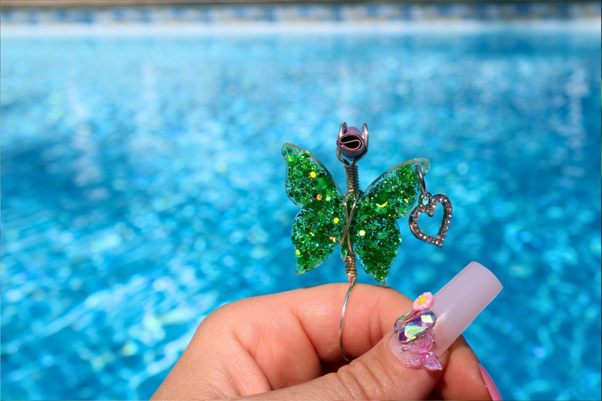 A manicured hand holds a whimsical, glittery green butterfly smoke ring with a heart charm in front of a sparkling swimming pool.