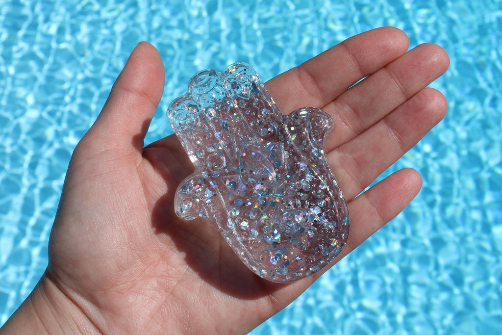 A hand holds a clear, glittery Hamsa hand against a vibrant blue pool background.