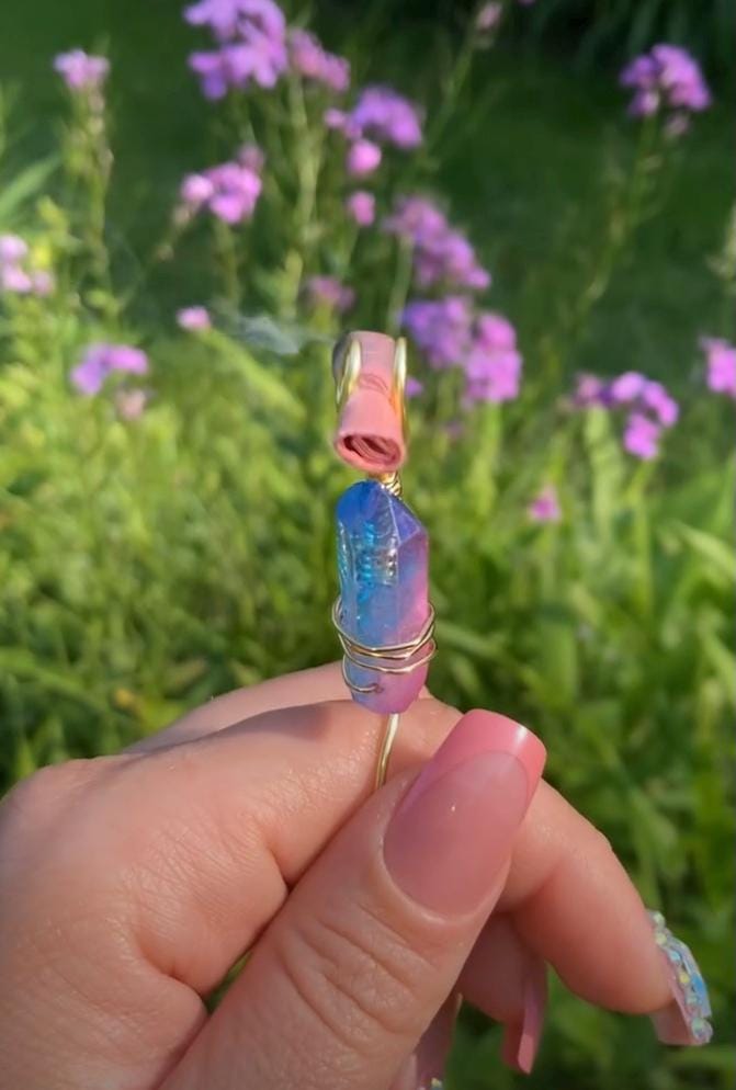A hand with pink nails holds a wire-wrapped aura crystal smoke ring against a background of purple flowers.