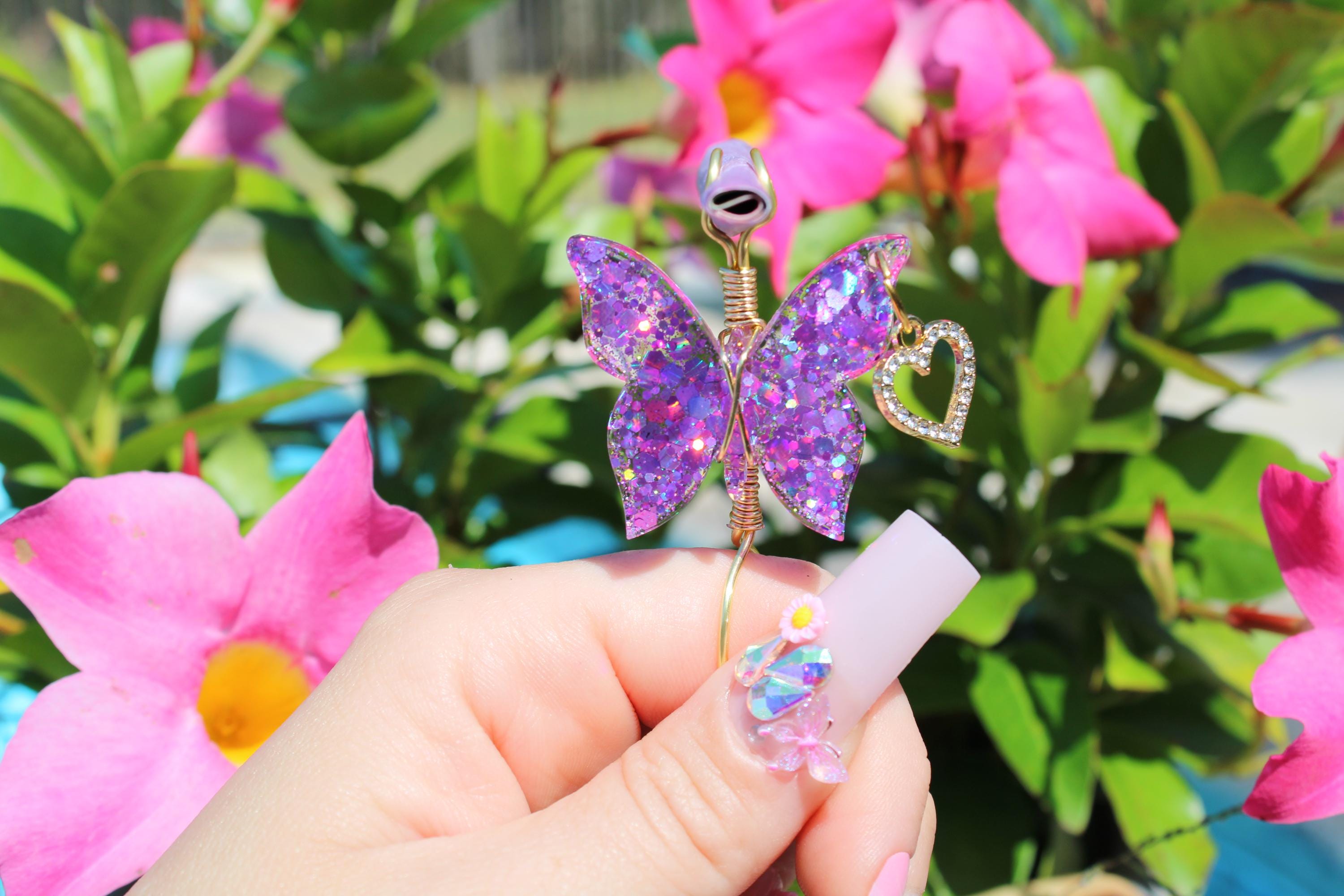 A hand with a decorative nail holds a handmade purple glitter butterfly smoke ring with a heart charm against a backdrop of bright pink flowers and green foliage.