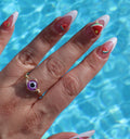 A woman's hand displays summer nail art with watermelon and strawberry decorations and a purple evil eye ring against a pool background.