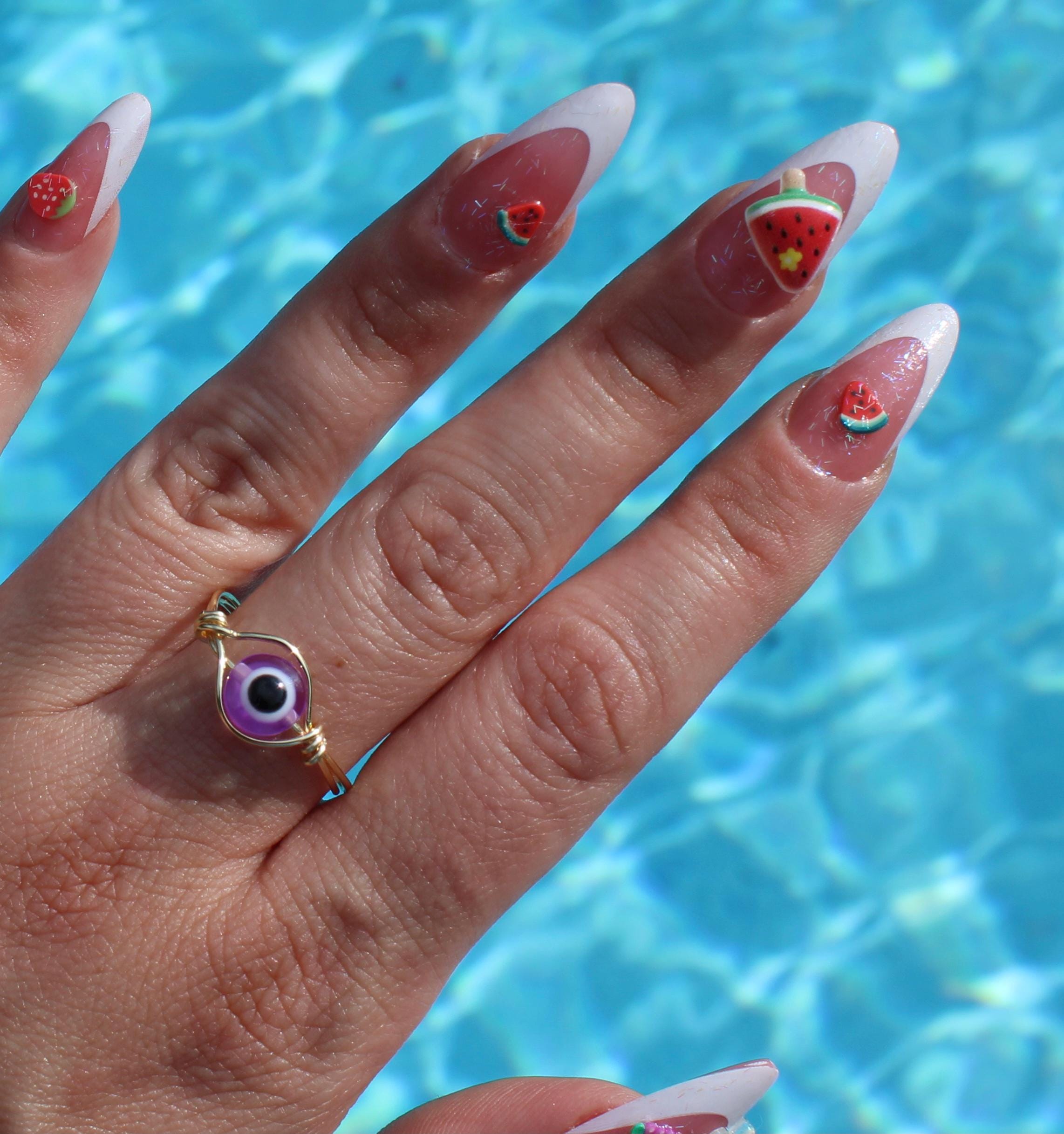 A woman's hand displays summer nail art with watermelon and strawberry decorations and a purple evil eye ring against a pool background.