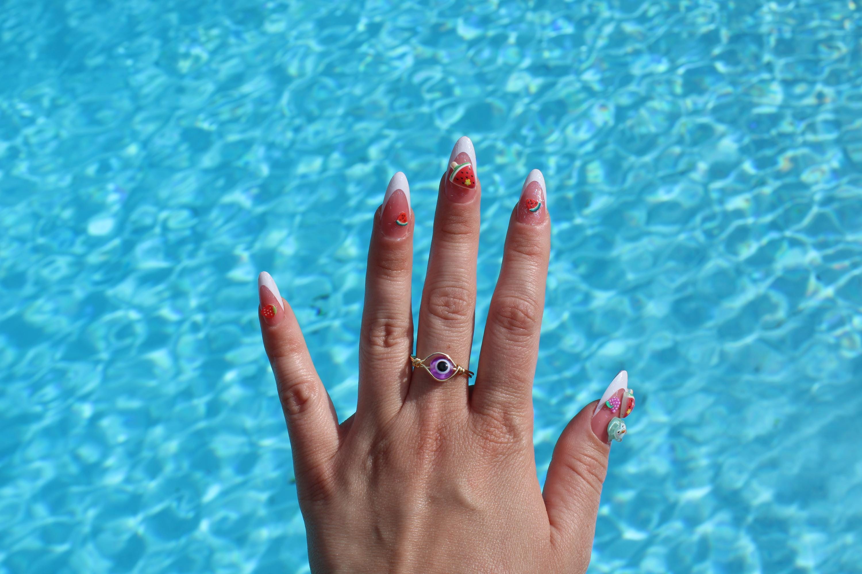 Woman shows off her trendy summer nails, featuring watermelon, strawberry nail art, and a purple evil eye ring, against a bright blue pool background.
