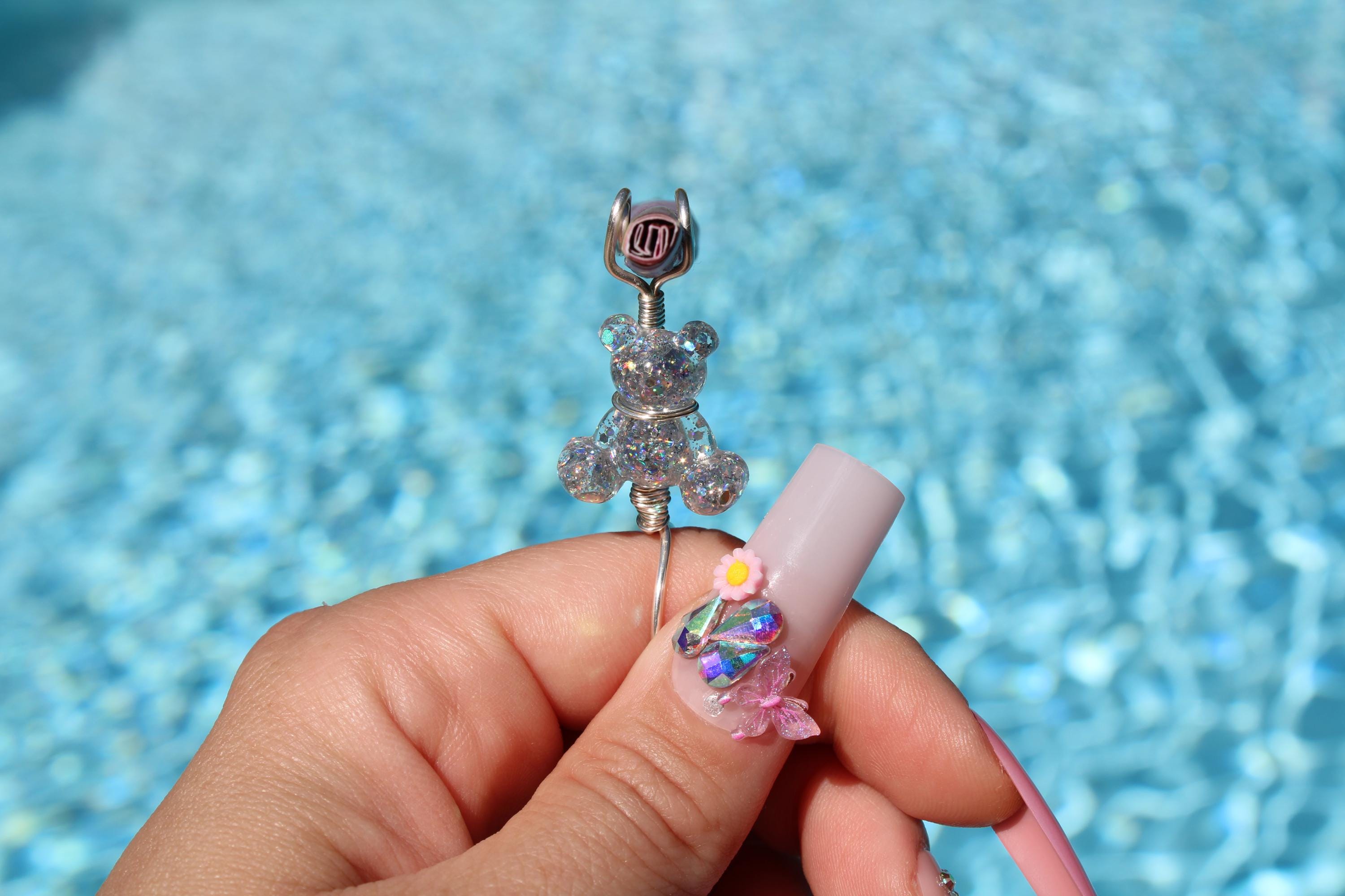 A hand with a decorated long pink nail holds a sparkling teddy bear smoke ring against a blurred swimming pool background.