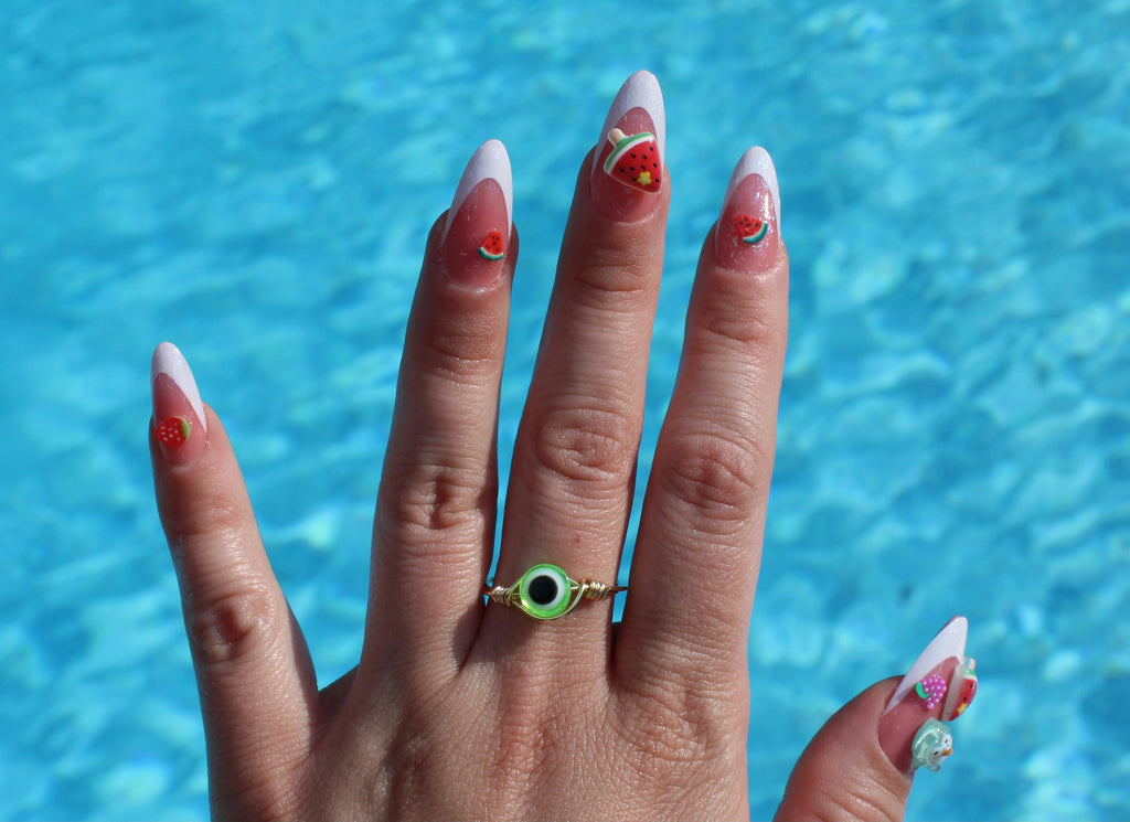 Hand with decorated nails and a green ring in front of a blue water background
