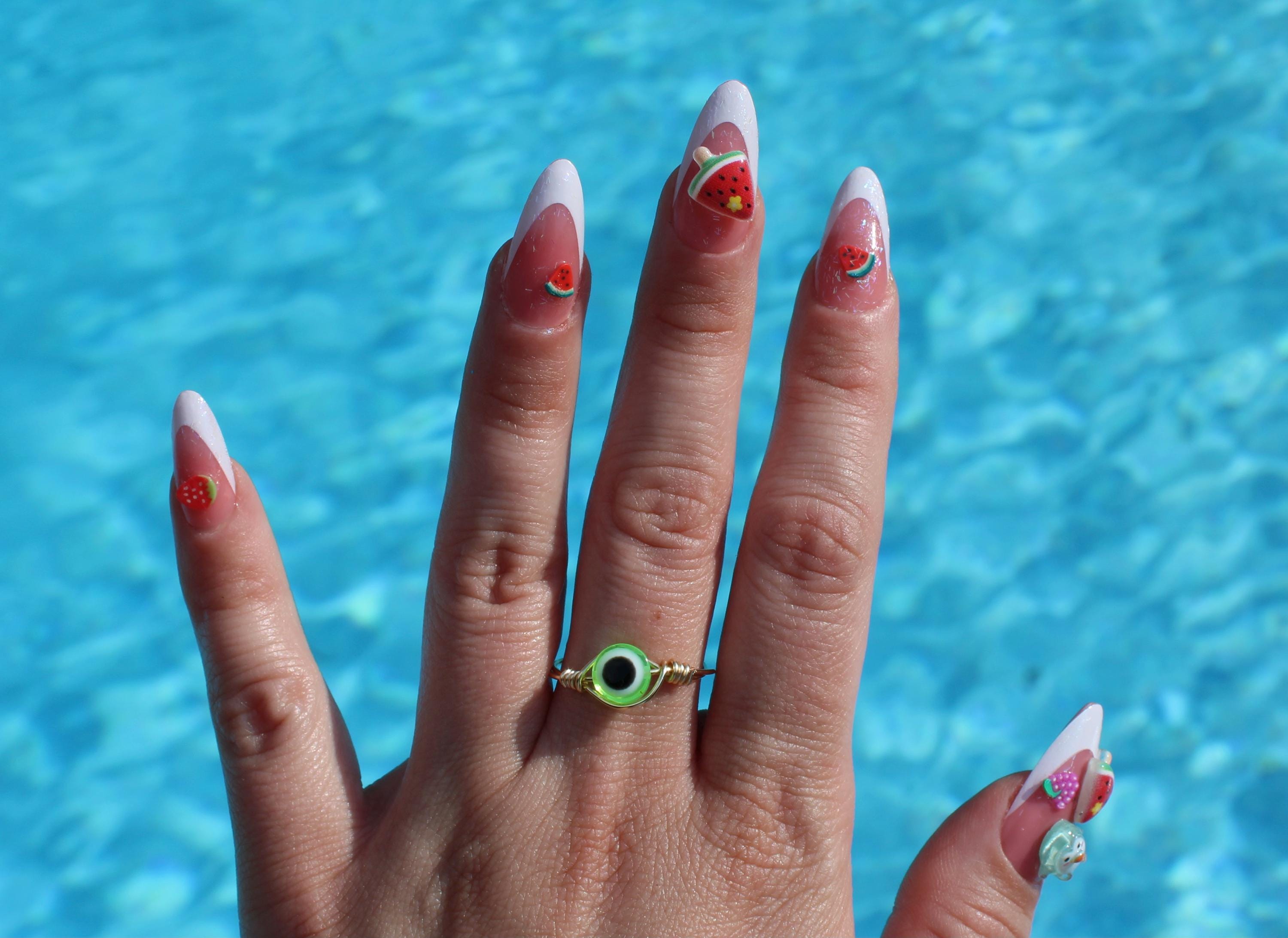 Hand with decorated nails and a green ring in front of a blue water background
