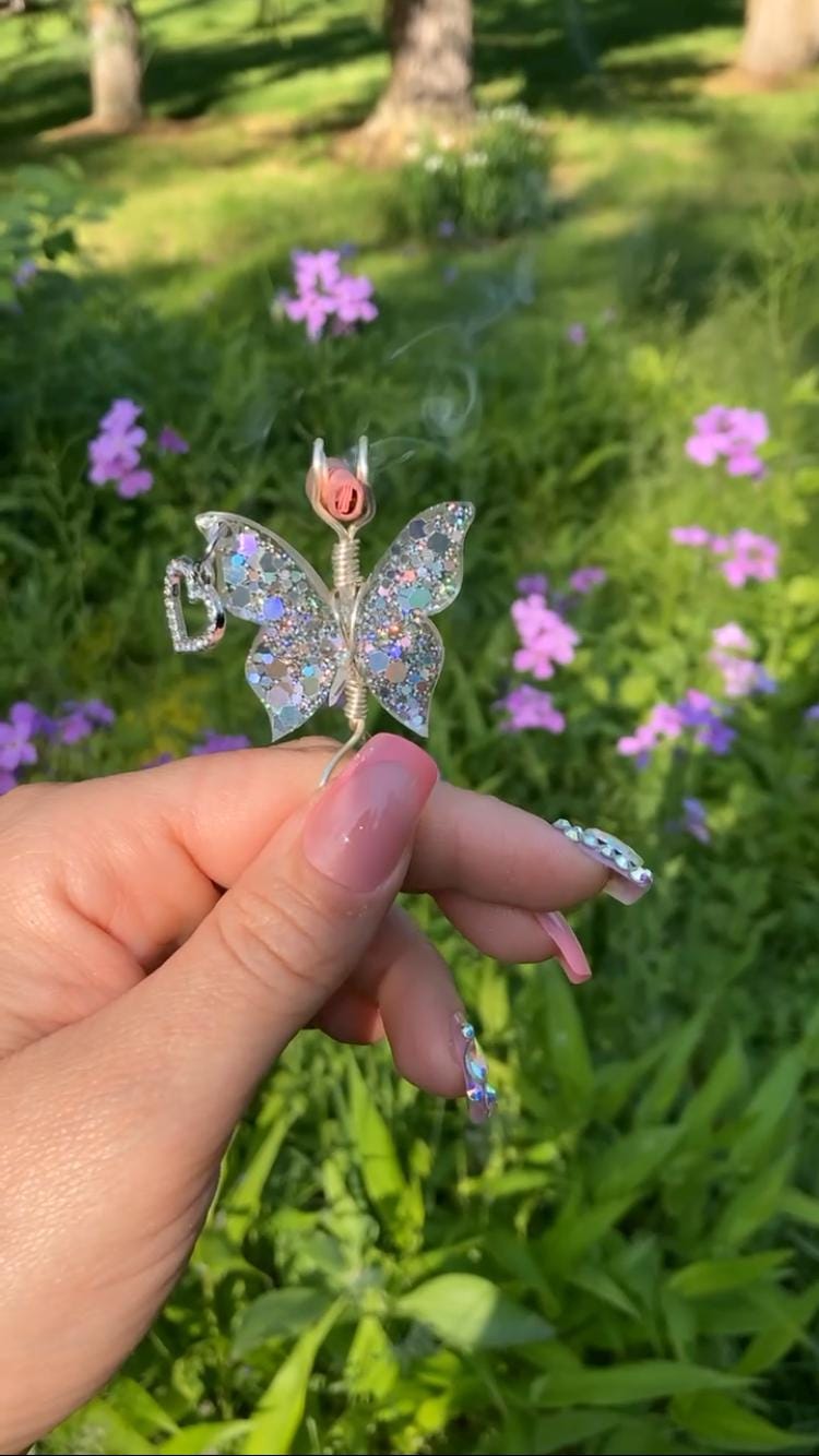 A hand with pink nails holds a sparkling, whimsical butterfly joint holder against a backdrop of purple flowers and greenery.