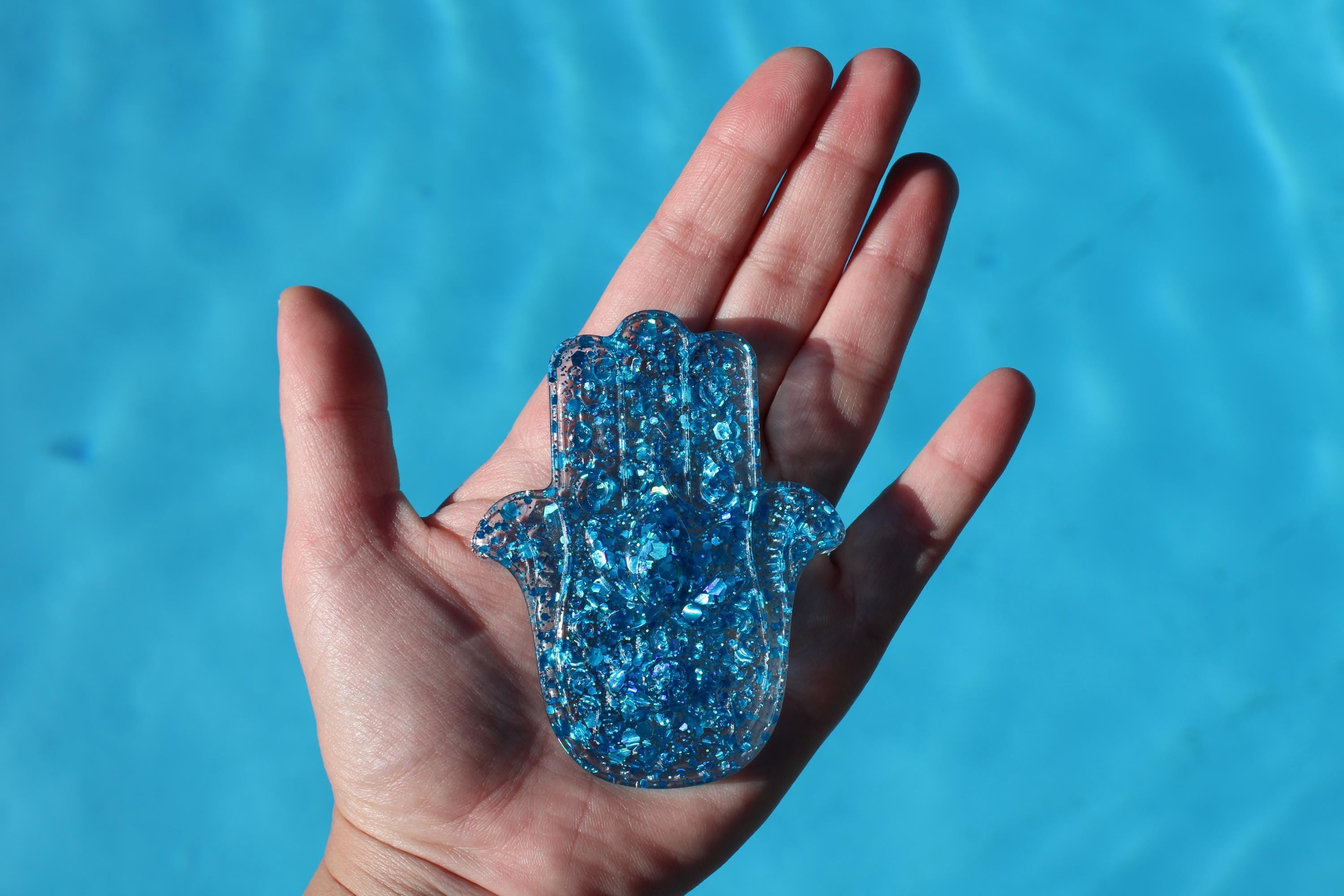 Blue glittery Hamsa hand held in a hand against a blue water background