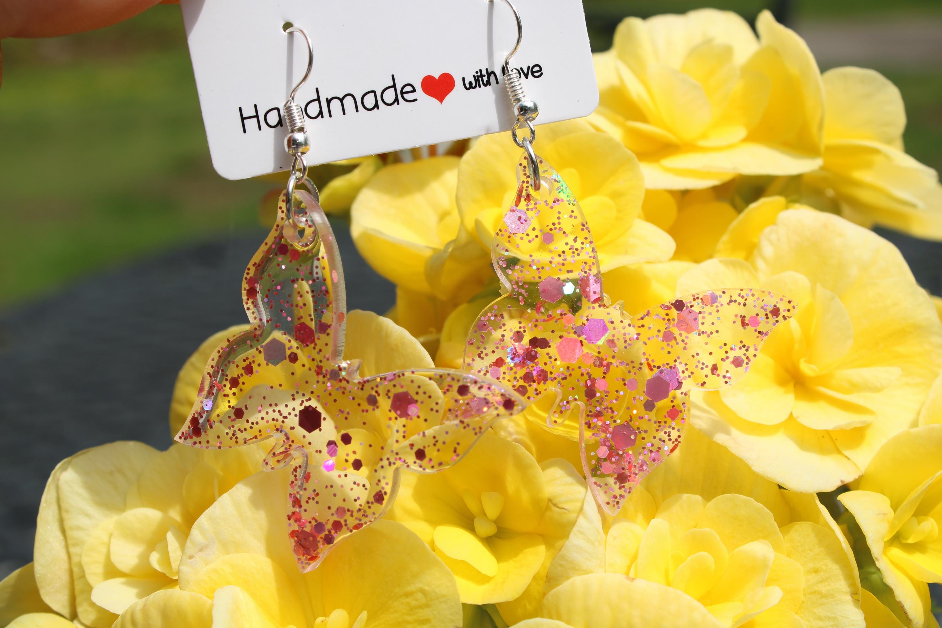 Butterfly-shaped earrings with glittery finish on a card labeled 'Handmade with love' against a yellow flower background.