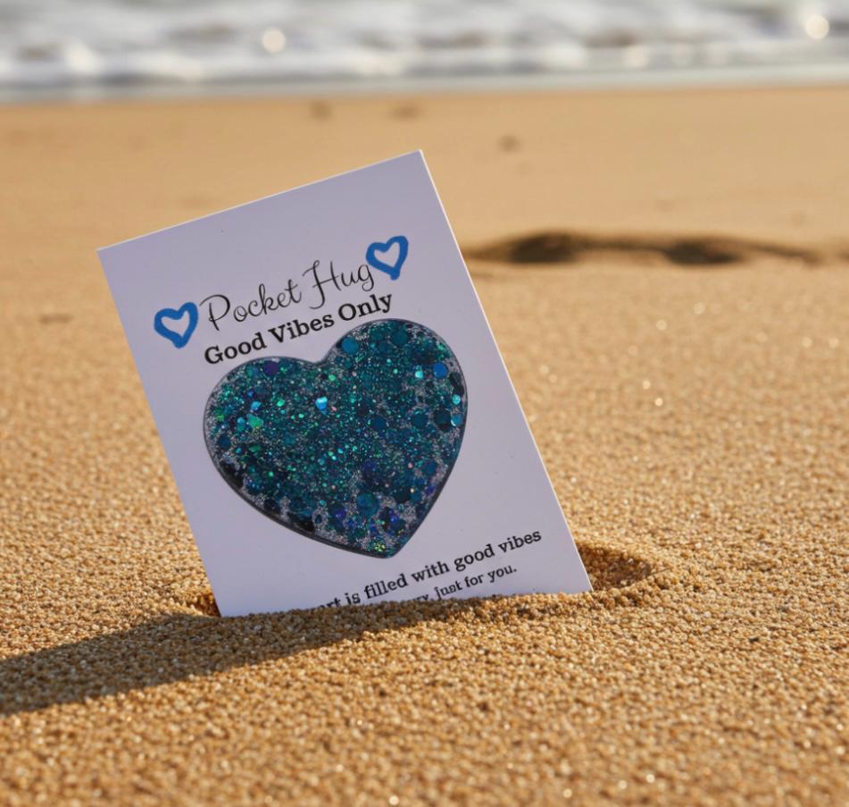 Heart-shaped glittery object on a 'Pocket Hug Good Vibes Only' card on sand