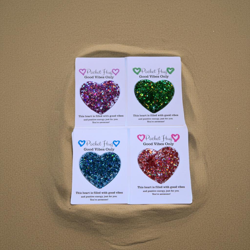 Four heart-shaped glittery objects on cards with 'Pocketful of Good Vibes Only' text on a beige background.
