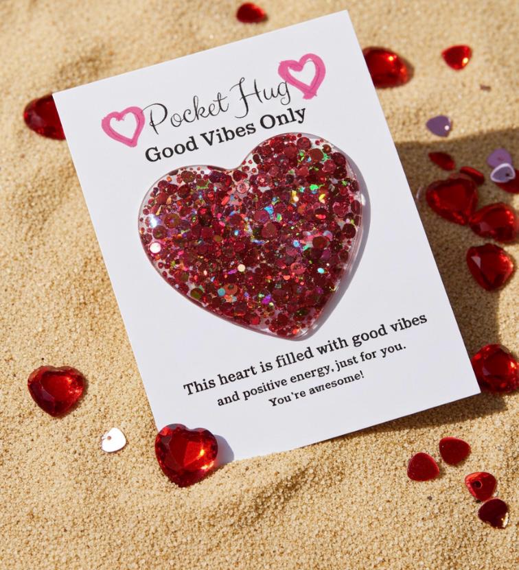 Heart-shaped glittery object on a card with 'Pocket Hug' text, surrounded by red hearts on sand.