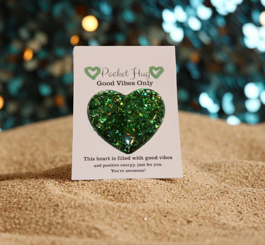 Green glitter heart on a card with 'Pocket Hug Good Vibes Only' text, against a blurred festive background.