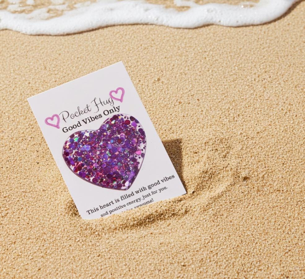 Heart-shaped glittery object on a card with 'Pocket Hug Good Vibes Only' text, lying on sand with ocean foam.