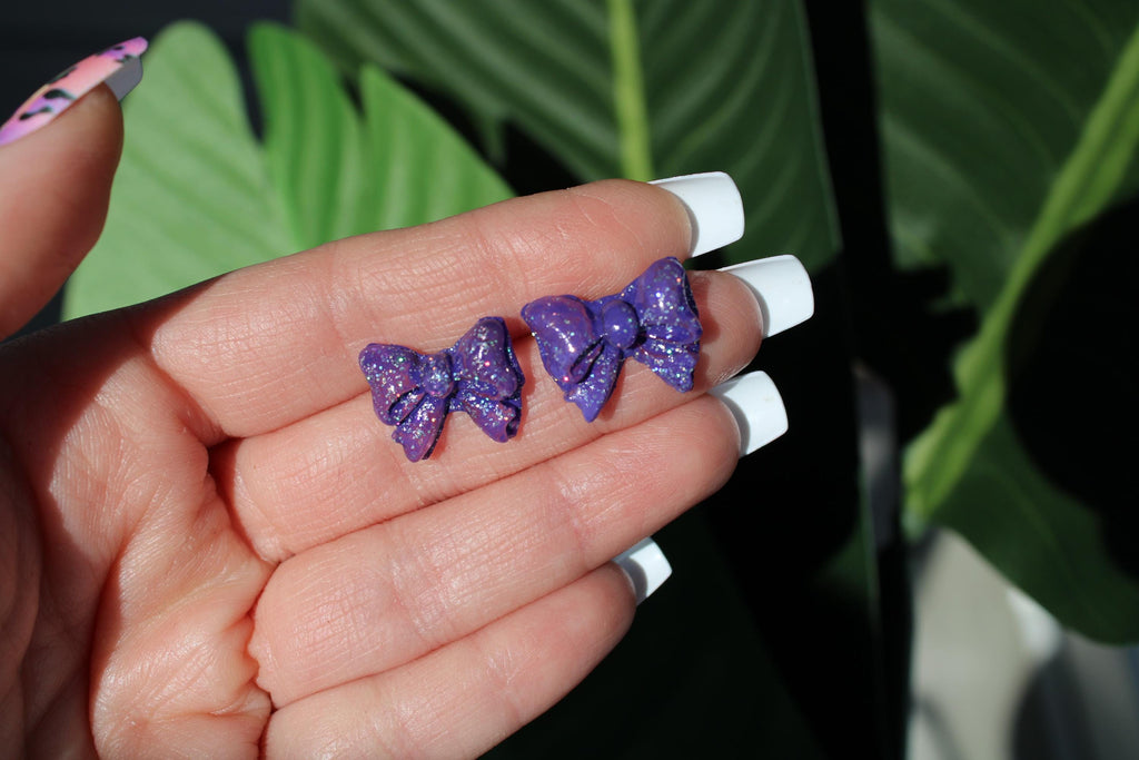 Purple glittery bow-shaped earrings held in a hand with a blurred green background