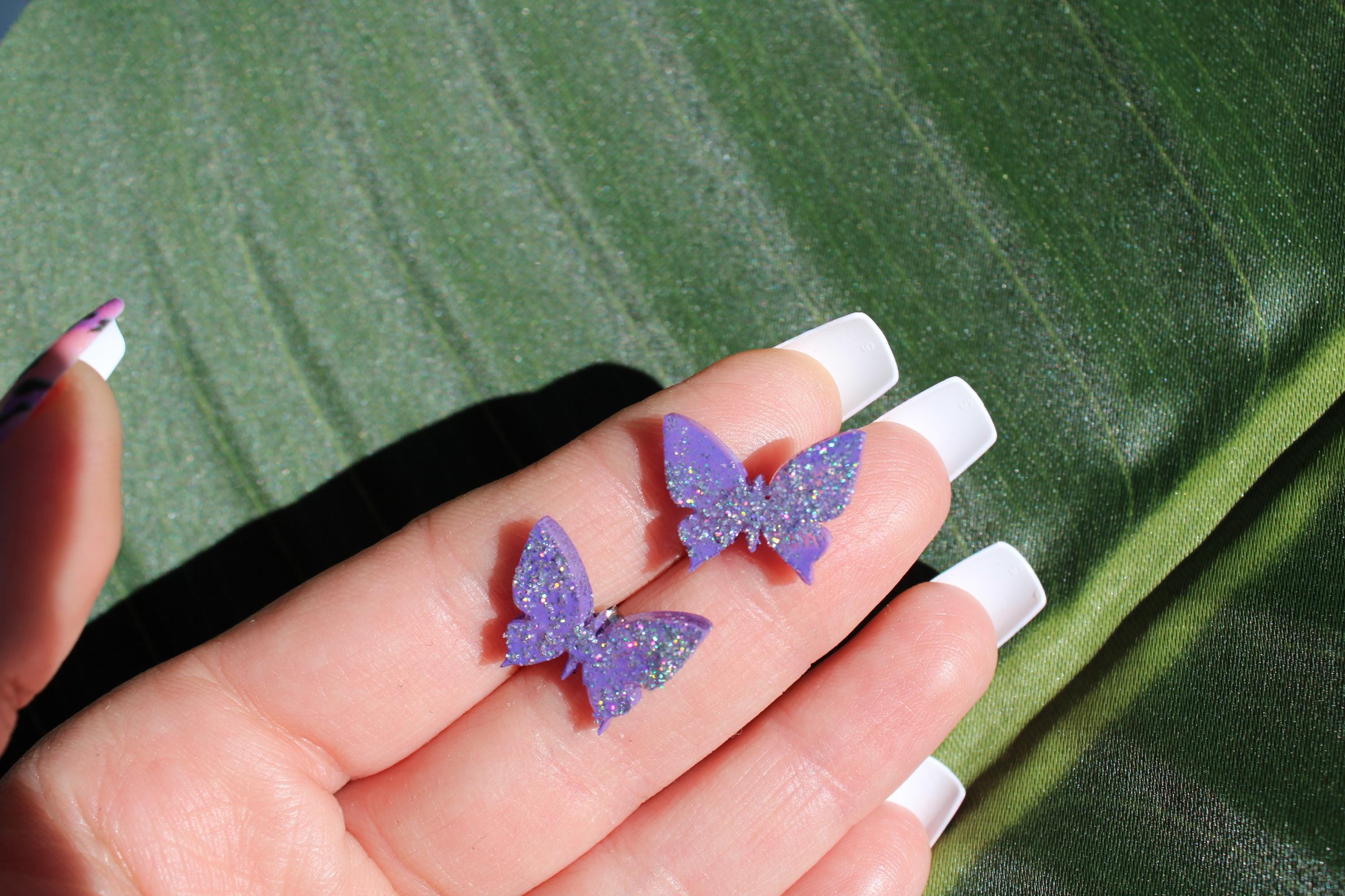 Purple butterfly earrings held between fingers against a green leaf background