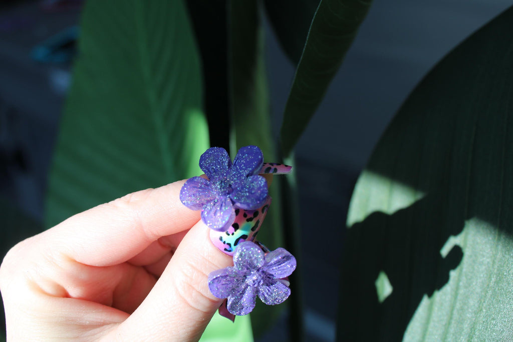 Hand holding a small purple flower with green leaves in the background
