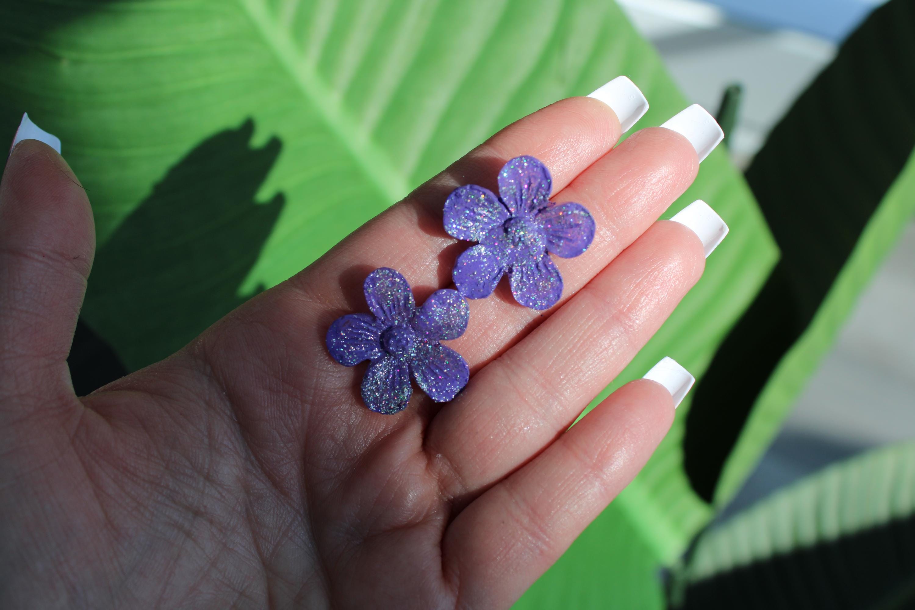 Hand holding two purple glittery flower-shaped objects with a green leafy background