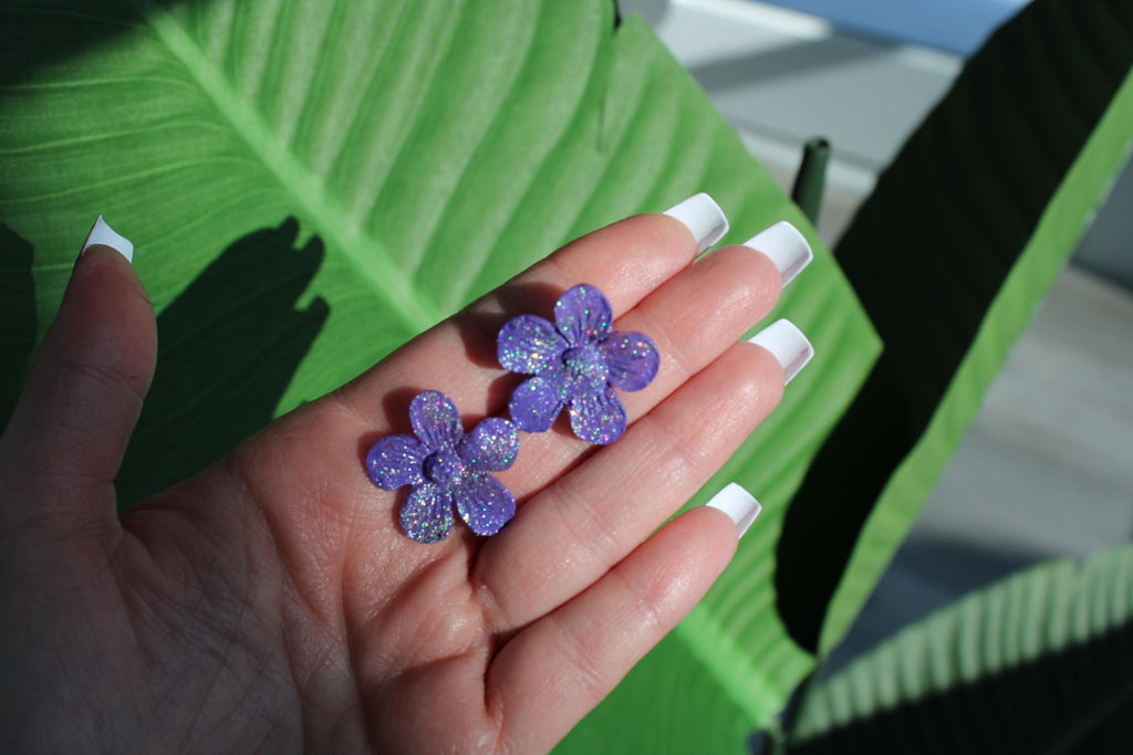 Hand holding two purple flower-shaped earrings with a green leafy background