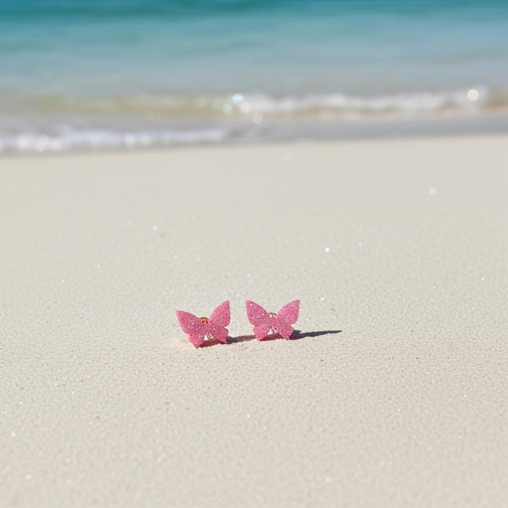 Pink butterfly earrings on a sandy beach with ocean waves in the background