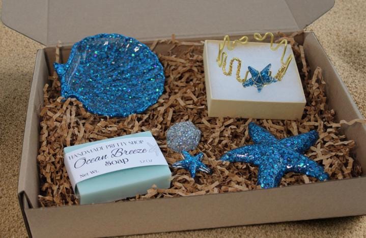 Box with decorative items including a blue glittery seashell soap dish, three resin shell art, a bar of soap and a starfish bracelet on a sandy background.