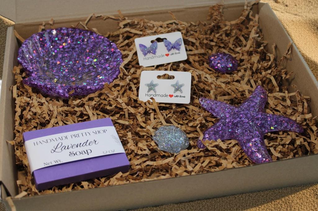 Box with purple glittery seashell resin dish, purple butterfly-shaped stud earrings, glitter starfish stud earrings, three resin art shells and lavender soap on brown shredded paper.