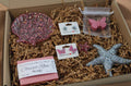 Box of rose scented handmade soap and matching mini butterfly rose scented soap, a resin starfish, pink butterfly stud earrings, and glitter starfish earrings on paper in a gift set.