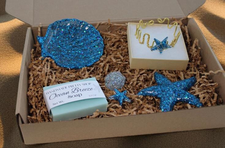 Box with decorative items including a blue glittery seashell soap dish, three resin shell art, a bar of soap and a starfish bracelet on a sandy background.