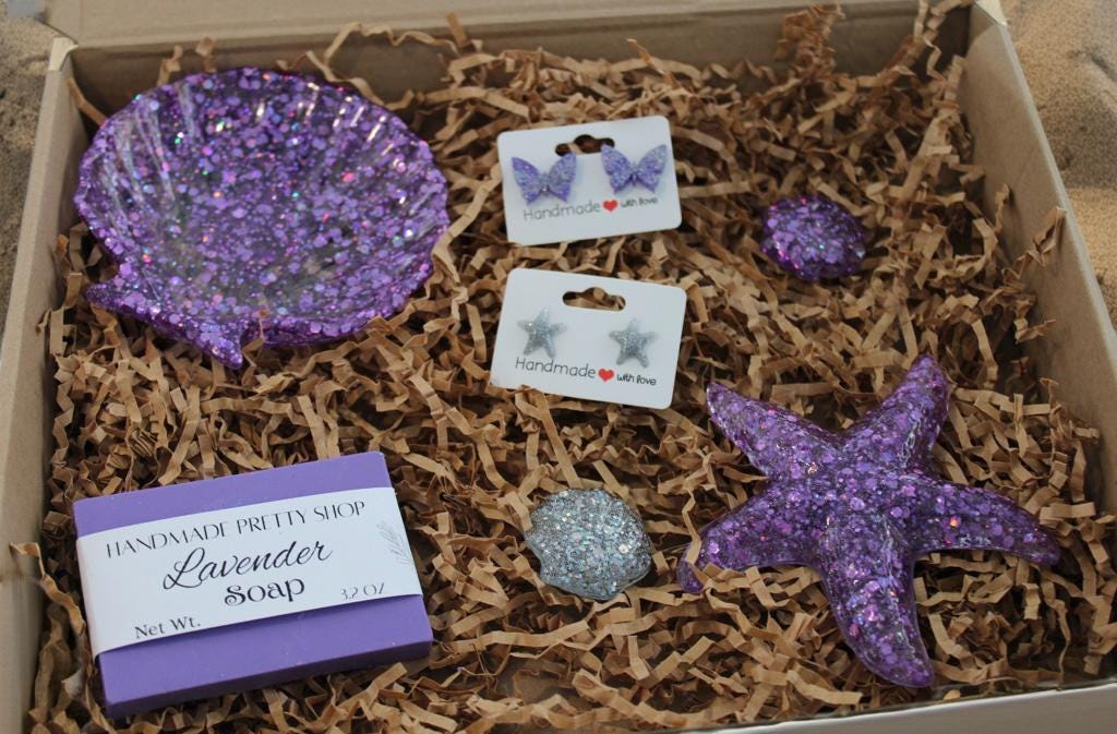 Box with purple glittery seashell resin dish, purple butterfly-shaped stud earrings, glitter starfish stud earrings, three resin art shells and lavender soap on brown shredded paper.