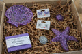 Box with purple glittery seashell resin dish, purple butterfly-shaped stud earrings, glitter starfish stud earrings, three resin art shells and lavender soap on brown shredded paper.