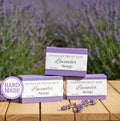 Three bars of lavender soap with 'Handmade Pretty Shop' branding on a wooden surface with a lavender field in the background.