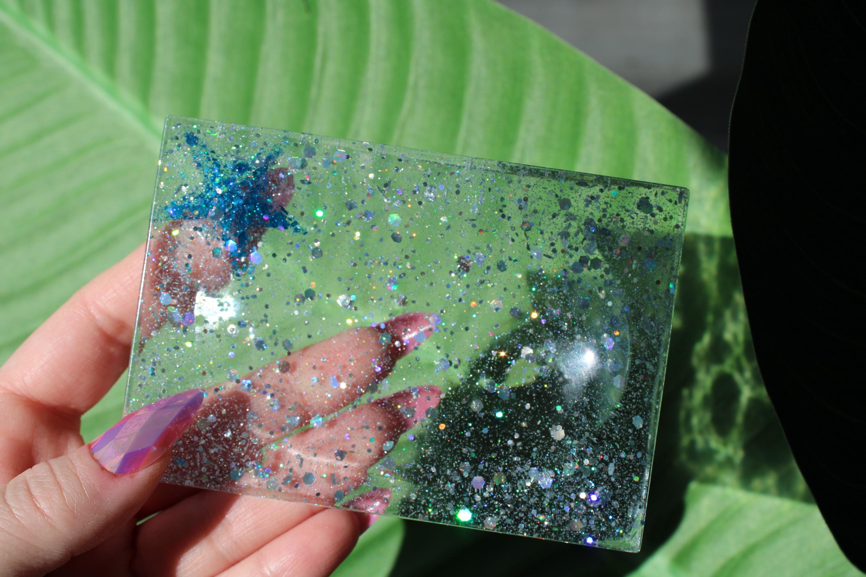 Hand holding a glittery square soap dish against a green leaf background