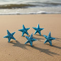 Five blue resin starfish on a sandy beach with ocean waves in the background.