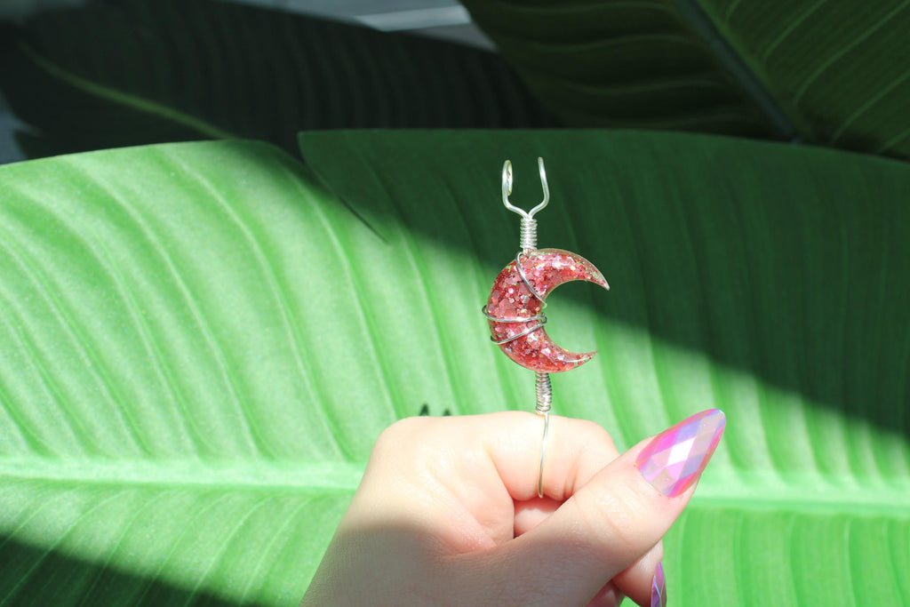 Hand holding a pink glittery crescent moon smoke ring against a green leaf background