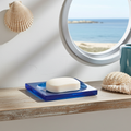 A bar of soap rests on a blue soap dish on a weathered shelf, evoking a coastal bathroom aesthetic with a seashell and ocean view visible through a porthole window.