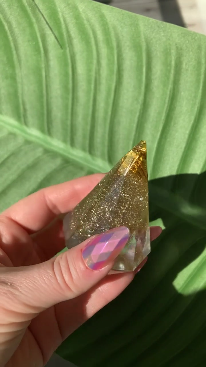 Video shows a resin ring display holder with gold glitter being held by a manicured hand in front of a leafy background 