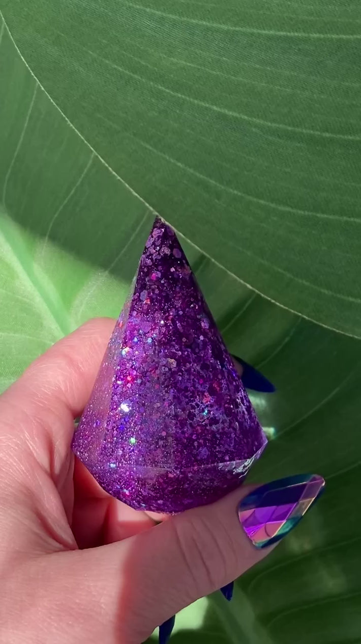 Video shows a manicured hand holding a purple sequin ring holder resin display in front of a leafy background 