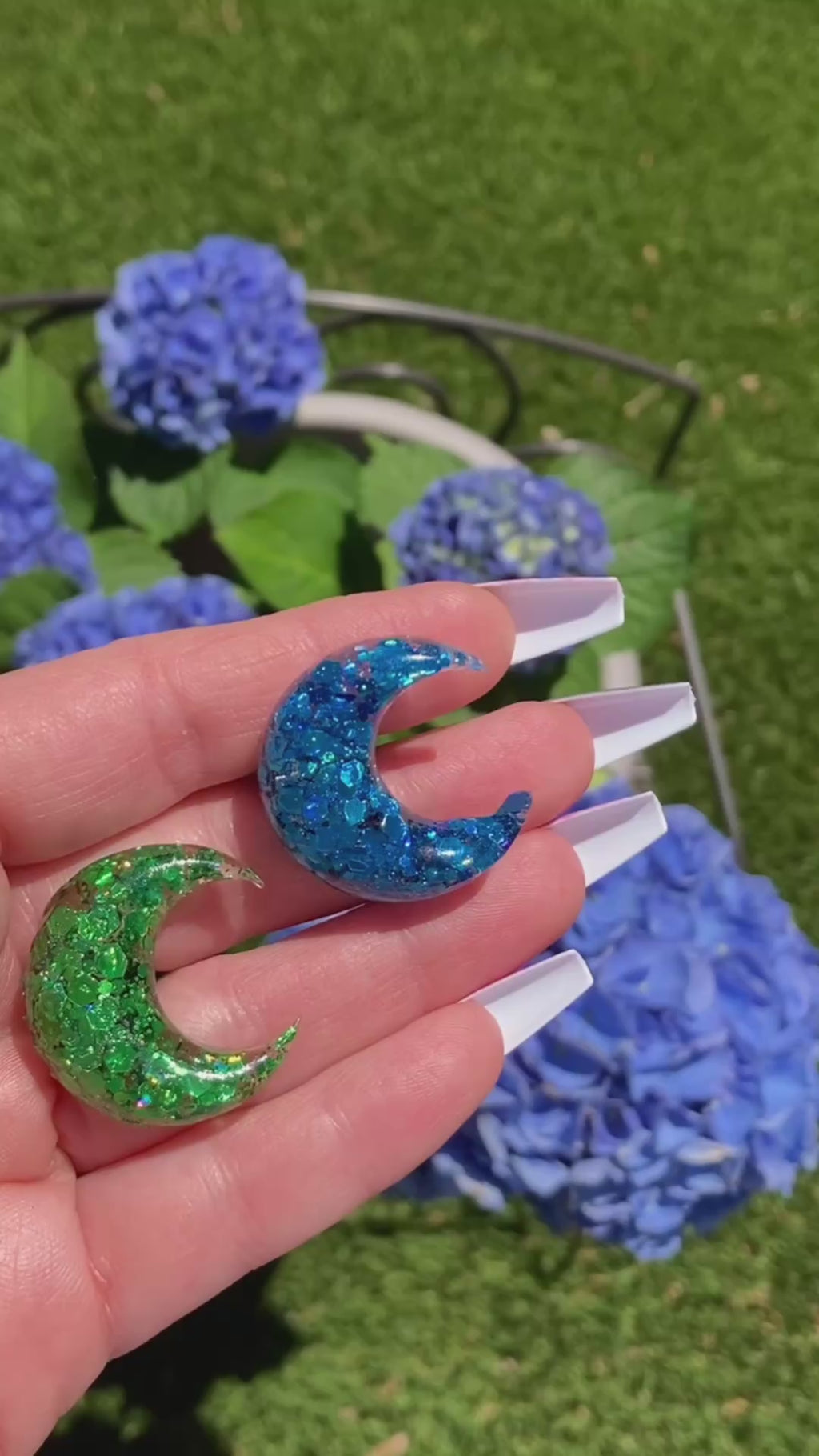 Video shows two moon car vent clips one green sequin and one blue sequin in a hand in front of flowers 